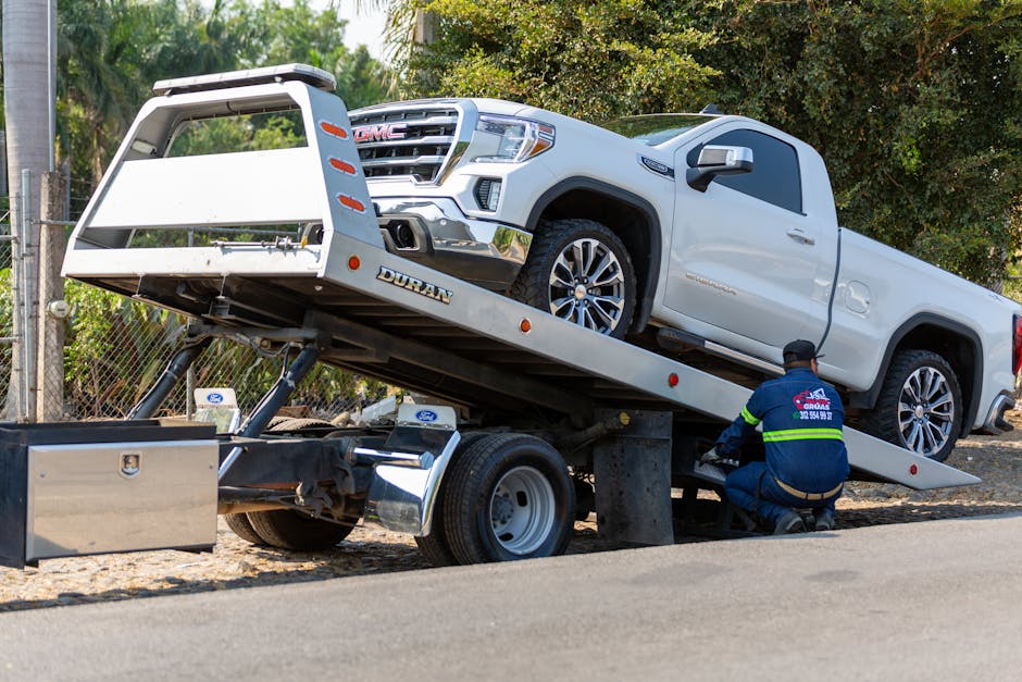 Tow truck operator in a hi-vis safety vest preparing a flatbed tow truck to load a white pickup